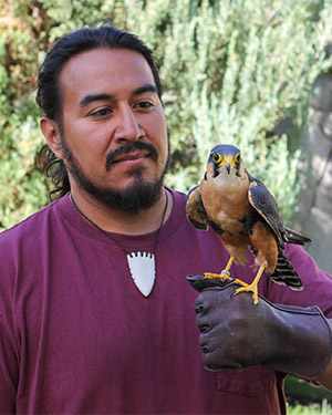 Arcia stoically poses with a kestrel. He is wearing a necklace and tshirt and has short facial hair. He has his hair tied back in a low bun.