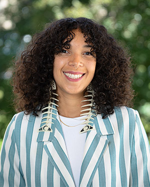 Airel smiles while wearing dramatic, long earrings and a striped blazer. She has shoulder-length hair in tight curls.