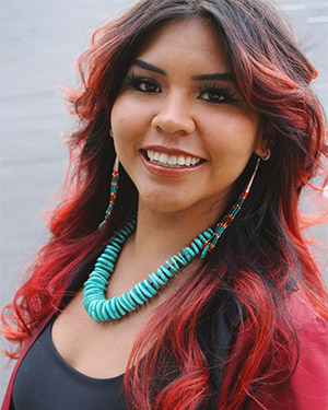 Jessica smiles while wearing turquoise beaded necklace and long earrings. She has her long hair styled in loose curls.