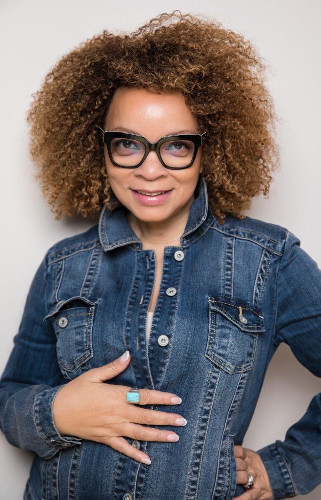 Ruth e. Carter smiles in a denim top, thick framed glasses, and has curly shoulder-length hair 