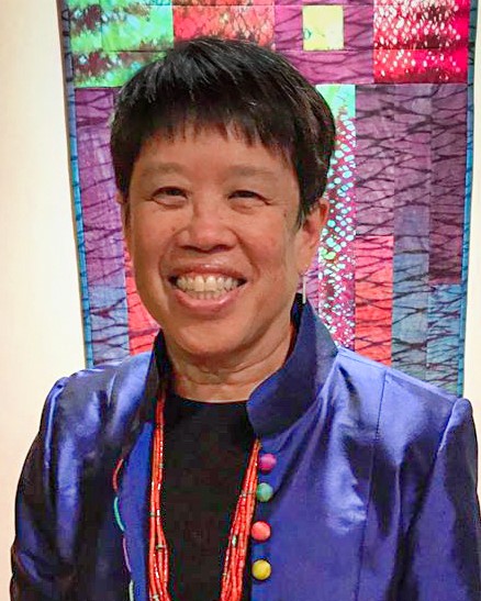 Marjorie A. Chan smiles in front of a textile art piece. She is wearing an open high-neck blazer and shirt. She has a short pixie cut hairstyle.