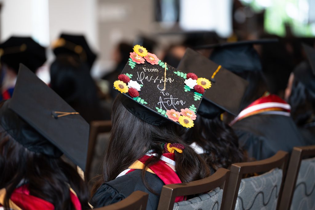 a row of students sitting in graduation regalia, one of their caps is decorated with flowers and a quote 'from passion to action'